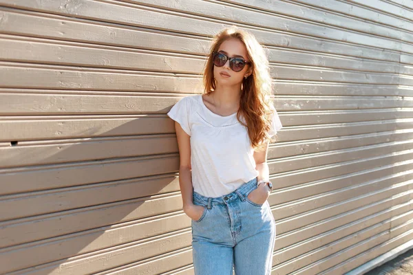 Jolie jeune femme hipster aux lunettes de soleil élégantes en t-shirt blanc tendance en jean vintage pose près d'un mur en bois par une journée d'été ensoleillée. modèle fille à la mode bénéficie du printemps — Photo