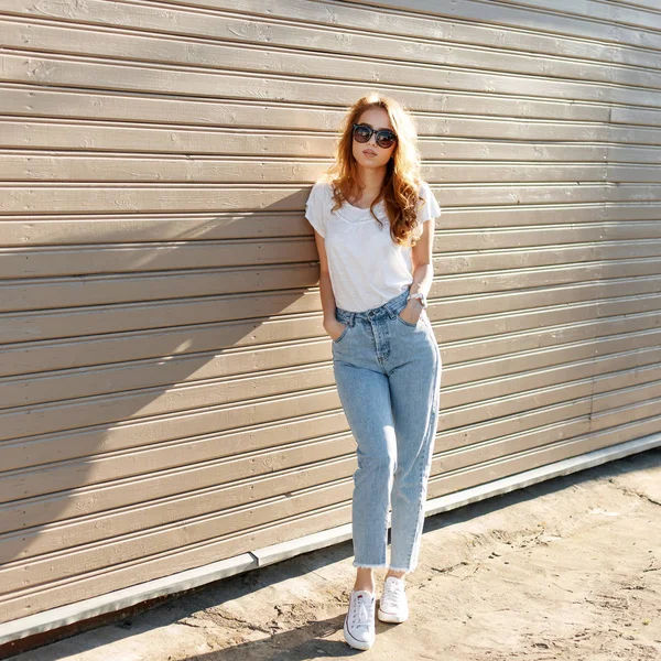 Europeiska ganska ung hipster kvinna i snygg vit T-shirt i vintage jeans i trendiga solglasögon stående nära trä väggen och njuter av sommaren. Söt flicka resor. — Stockfoto