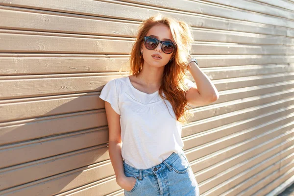 Stylish beautiful young model woman with sunglasses in a white T-shirt and blue vintage jeans near a wooden wall at sunset. Fashion hipster summer season — Stock Photo, Image