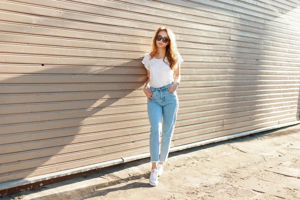 Glamouröse hübsche junge Hipsterfrau im sommerweißen T-Shirt in Turnschuhen in trendigen Jeans mit stylischer Sonnenbrille steht an einer Holzwand in der Stadt. europäisches Mädchen bei einem Spaziergang an einem sonnigen Sommertag — Stockfoto