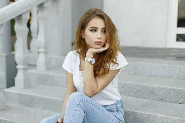 Élégante jeune jolie femme dans un t-shirt blanc à la mode en jeans bleu à la mode se trouve sur les marches vintage de l'hôtel à l'extérieur. Américain attrayant fille modèle se détend par une journée chaude d'été . — Photo