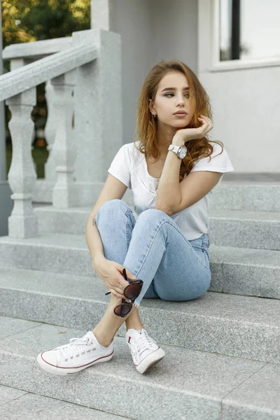 Młoda ładna kobieta w stylowej koszulce w dżinsy w modne trampki odpoczywa, siedząc na schodach budynku vintage. Ładna dziewczyna model relaksuje się na zewnątrz. Letni styl Odzież damska. — Zdjęcie stockowe