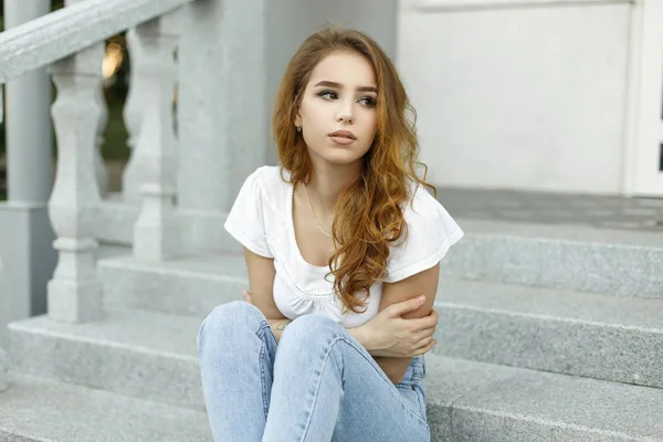 La joven europea bonita de moda en una camiseta blanca con estilo en jeans azules de moda está descansando en la escalera vintage al aire libre. Bonita chica modelo se sienta y sueña con un cálido día de primavera . — Foto de Stock