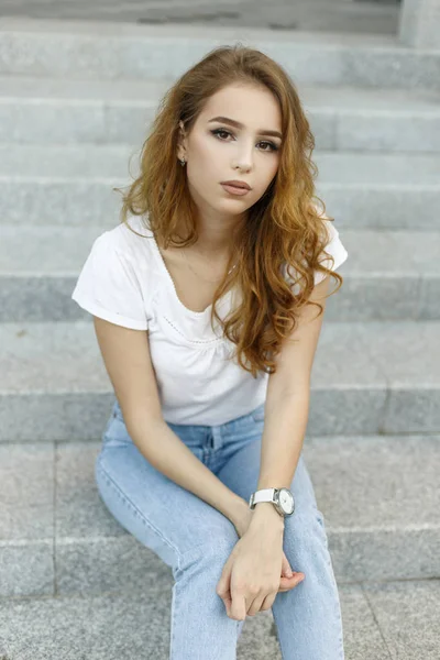 Jovem mulher bonita e elegante em jeans vintage na moda em uma camiseta elegante branca está descansando sentado nos degraus. Menina de moda bonita americana em férias . — Fotografia de Stock