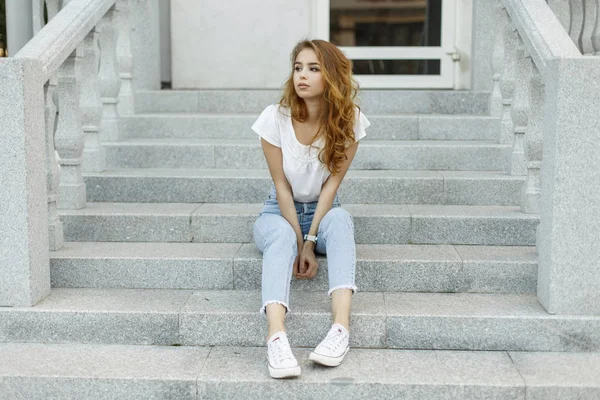 빈티지 건물의 계단에 앉아 휴식 하는 흰색 유행 스 니 커 즈에 청바지에 유행 t-셔츠에 매력적인 세련 된 젊은 여자. 여름 하루를 즐기는 귀여운 유럽 여자. — 스톡 사진