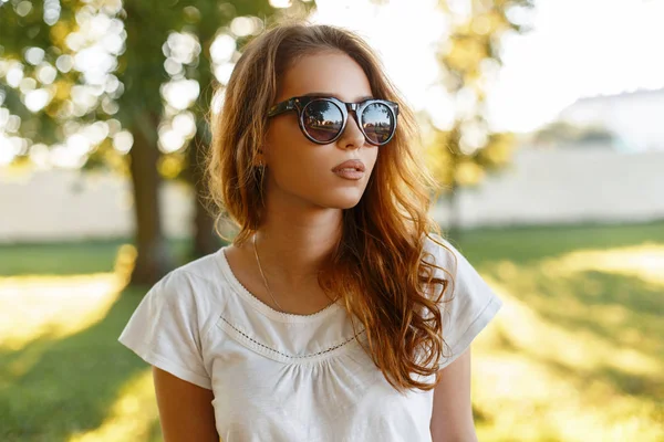 Portret kobiety hipster amerykański cute młoda redhead z sexy usta w stylowych okularów przeciwsłonecznych w modny, biały t-shirt w parku na tle jasny lato pomarańczowy zachód słońca. Ładna dziewczyna. — Zdjęcie stockowe