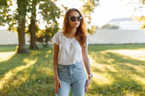 Европейская стильная молодая хипстерша в модных джинсах в белой футболке в модных солнцезащитных очках позирует в парке среди зеленых деревьев на фоне летнего заката. Современная симпатичная девочка любит ходить . — стоковое фото