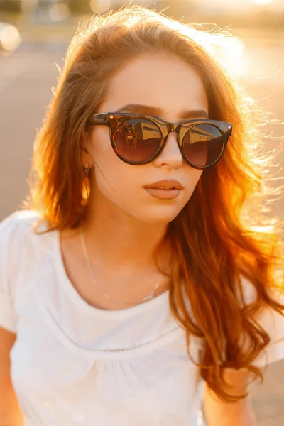 Retrato facial de una hermosa joven pelirroja hipster con elegantes gafas de sol en una camiseta blanca de moda en un parque de atracciones sobre un fondo de brillante puesta de sol de verano naranja. Niza chica moderna . —  Fotos de Stock