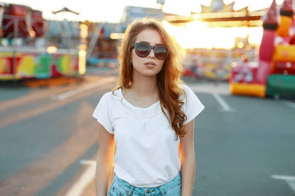 Attrayant jeune femme hipster élégant avec des cheveux roux dans des lunettes de soleil dans un T-shirt blanc en jeans se promène dans un parc d'attractions par une chaude journée ensoleillée de printemps. Jolie fille profiter du week-end . — Photo