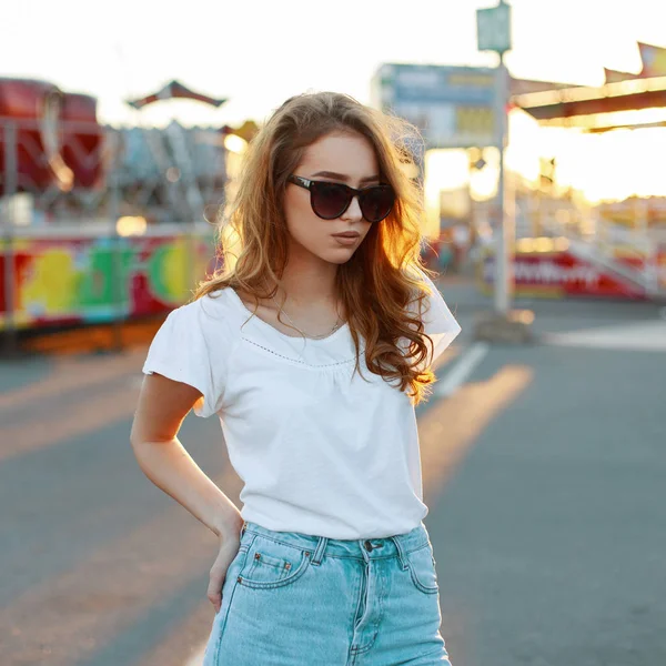 Руда модна молода хіпстерка в білій футболці в стильних сонцезахисних окулярах в блакитних вінтажних джинсах відпочиває в парку розваг на фоні заходу сонця. Приваблива дівчина у відпустці . — стокове фото