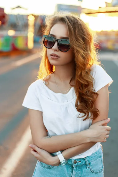 Élégante rousse américaine jeune femme hipster en lunettes de soleil tendance dans un t-shirt en jean vintage posant sur le fond des carrousels dans le parc par une chaude journée ensoleillée. Fille mignonne aime le week-end . — Photo