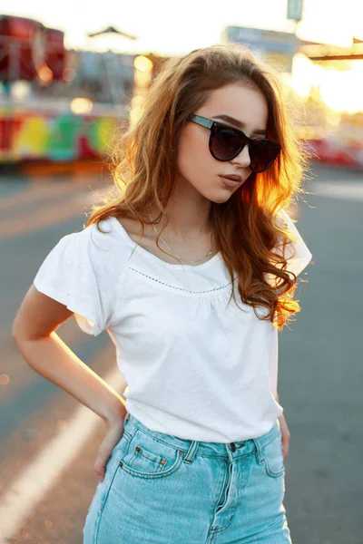 Carino bella rossa giovane hipster donna in occhiali da sole alla moda in una t-shirt in jeans alla moda in un parco divertimenti sullo sfondo di un tramonto estivo luminoso. Bella ragazza modello su una passeggiata . — Foto Stock