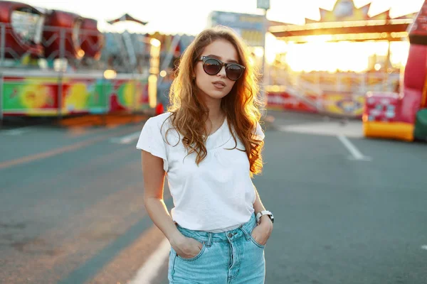 Jovem mulher hipster elegante em jeans na moda em óculos de sol elegantes na moda t-shirt está desfrutando do sol brilhante verão em um parque de diversões. Menina bonita modelo de moda descansando ao ar livre . — Fotografia de Stock