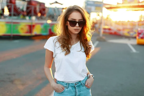 Bonito muito elegante mulher jovem hipster em jeans vintage em óculos de sol na moda em uma camiseta branca elegante caminha e desfruta de um dia ensolarado de verão em um parque de diversões. Modelo menina agradável em férias . — Fotografia de Stock