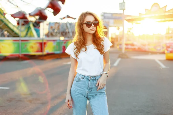 Όμορφη κοκκινομάλλα όμορφη νεαρή hipster γυναίκα σε κομψό μπλουζάκι σε vintage τζιν σε μοντέρνα γυαλιά ηλίου είναι το περπάτημα σε ένα λούνα παρκ. Μόδα χαριτωμένο μοντέλο κορίτσι απολαμβάνει ένα πορτοκαλί καλοκαιρινό ηλιοβασίλεμα. — Φωτογραφία Αρχείου