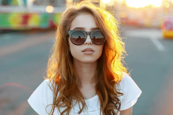 Portrait du visage d'une jeune jolie femme hipster dans des lunettes de soleil élégantes avec des cheveux bouclés rouges dans un t-shirt élégant sur le fond de soleil d'été orange vif. Fille élégante profiter de la lumière du soleil d'été . — Photo