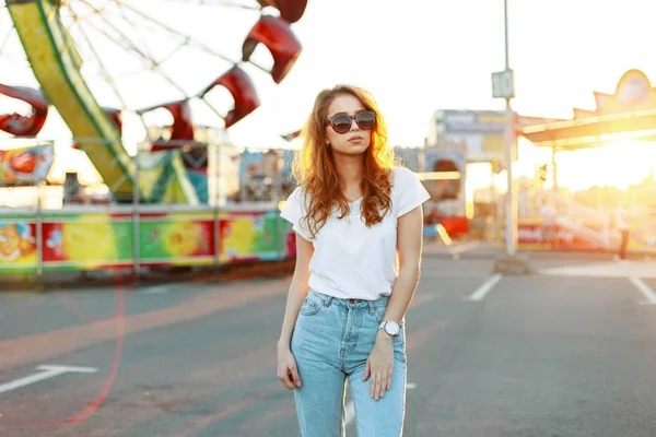 Ruiva bonita jovem mulher hipster bonito em elegante camiseta em jeans elegantes em óculos de sol da moda está andando em um parque de diversões. Menina modelo de moda incrível goza no pôr do sol de verão laranja . — Fotografia de Stock