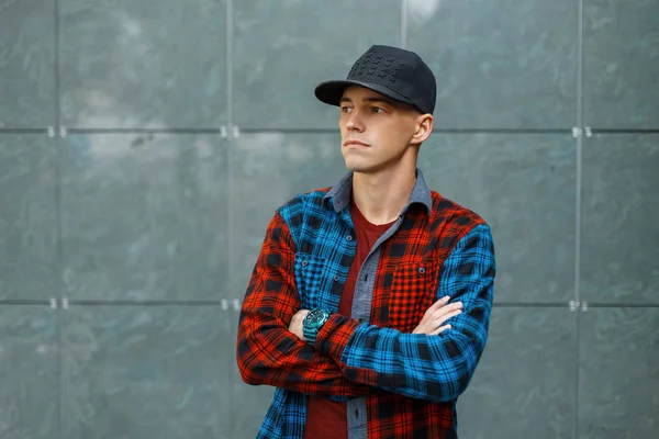 Guapo joven con estilo en una elegante camiseta en una camisa a cuadros azul y rojo de moda en una gorra de baloncesto de moda negro se encuentra cerca de una pared vintage gris. Un tipo genial. Ropa de hombre de estilo americano . — Foto de Stock