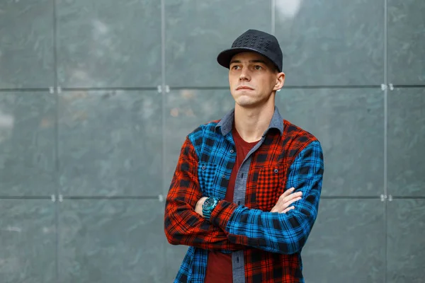 Hombre joven atractivo y elegante americano en una camiseta de moda en una camisa a cuadros azul y rojo elegante en una gorra de baloncesto de moda negro posa cerca de una pared vintage gris. Cool chico de moda . — Foto de Stock