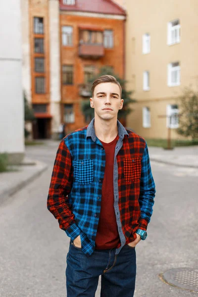 Young stylish handsome man in a red fashionable t-shirt in a blue and red plaid shirt walks through a small town on the background of old buildings. Pretty American guy is enjoying the weekend. — Stock Photo, Image