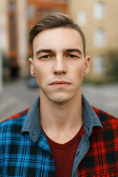 Young serious man with a stylish hairstyle in a fashionable t-shirt in a trendy plaid shirt walks around the city near the old brick buildings on a summer day. Modern guy. Portrait of a man's face. — Stock Photo, Image