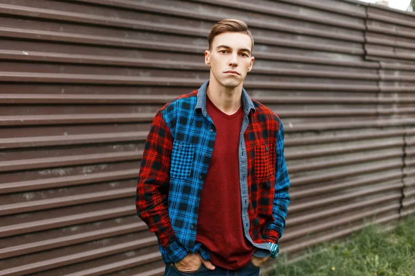 ブルー ジーンズ ビンテージ金属フェンス近く屋外でトレンディな格子縞のマルチカラー シャツでおしゃれな赤い t シャツでスタイリッシュなハンサムな若い男。魅力的なアメリカ人の男の散歩が好きです。. — ストック写真