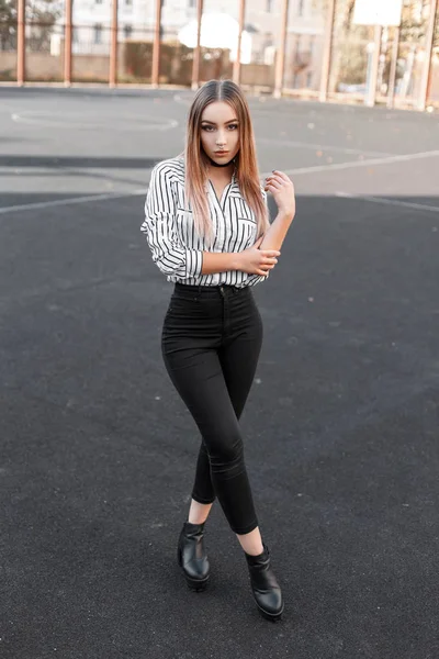 Glamorous modern young woman in black stylish jeans in a striped elegant blouse in trendy leather shoes posing on an open basketball court on a spring sunny day. Cute beautiful girl walks. — Stock Photo, Image