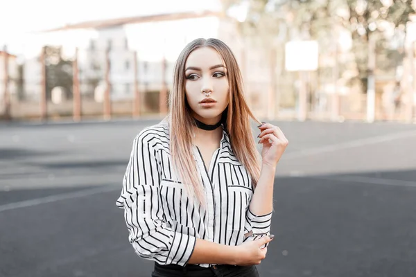 Linda joven adolescente con una nariz perforada en una camisa a rayas de moda en vaqueros negros con un collar de terciopelo negro se destaca al aire libre en un día soleado de primavera. Bastante modelo de chica moderna en el paseo —  Fotos de Stock