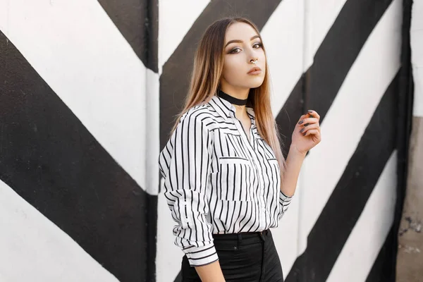 Mooie jonge vrouw in een trendy zwart-wit shirt in een lijn met een doordrongen neus in zwarte jeans in een halsketting van fluwelen poseren in de buurt van een gestreepte vintage muur. Glamour sexy meisje model op een wandeling. — Stockfoto