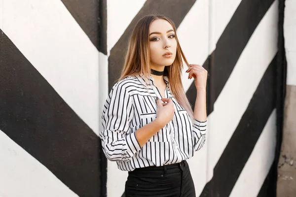 Modern pretty young woman in a trendy black and white blouse in a line with a pierced nose in black jeans in a vintage velvet necklace posing near a striped vintage wall. Cute girl fashion model. — Stock Photo, Image