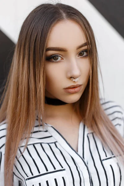 Portret van een vrij glamoureuze jongedame met mooie make-up met een trendy kapsel met een oorbel in haar neus in een stijlvolle gestreepte blouse met een fluweelzwart ketting in de buurt van een vintage muur. — Stockfoto