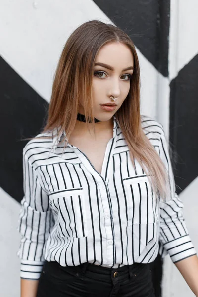 Mujer joven y bonita con una camisa blanca y negra de moda en una línea con una nariz perforada en jeans elegantes en una gargantilla de terciopelo se encuentra en la ciudad cerca de una pared vintage a rayas. Adolescente moderna en un paseo . —  Fotos de Stock