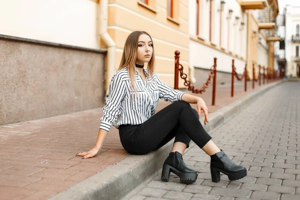 Schöne hübsche junge Frau in einer eleganten schwarz-weiß gestreiften Bluse in trendigen Jeans in Lederschuhen mit durchbohrter Nase sitzt draußen in der Stadt. Stilvolles Mädchen genießt das Wochenende. — Stockfoto