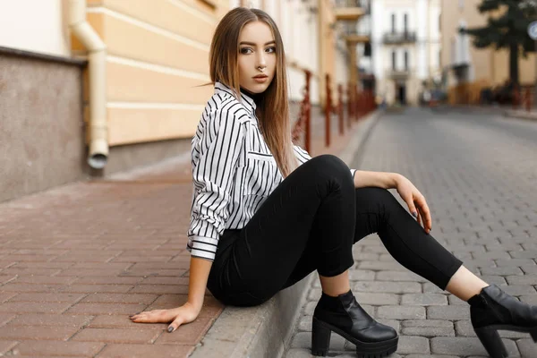 Jovem atraente moderna em jeans pretos em uma elegante blusa listrada em sapatos de couro está sentado em uma rua em um dia quente de primavera perto de edifícios vintage. Menina bonita relaxa na cidade . — Fotografia de Stock