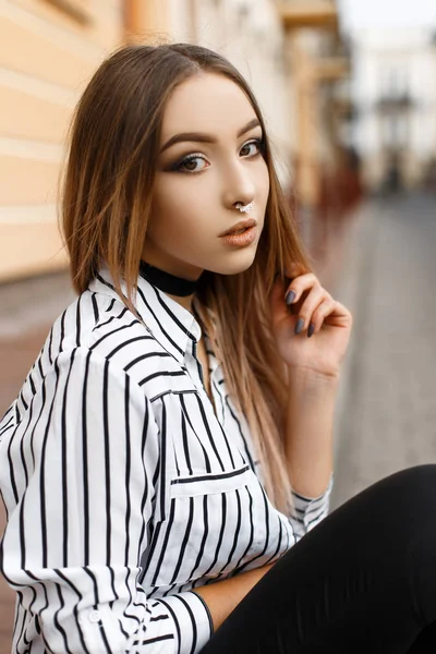 Bela jovem atraente em um colar de veludo com um brinco no nariz em uma blusa listrada elegante em jeans preto perto de um edifício vintage. Menina adolescente moderna na moda ao ar livre . — Fotografia de Stock