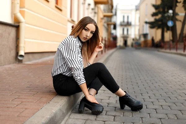 Coole hübsche junge Frau in eleganter schwarz-weiß gestreifter Bluse in trendigen Jeans in schwarzen Lederschuhen mit gepiercter Nase, die draußen in der Stadt sitzt. Modernes Mädchen genießt das Wochenende. — Stockfoto