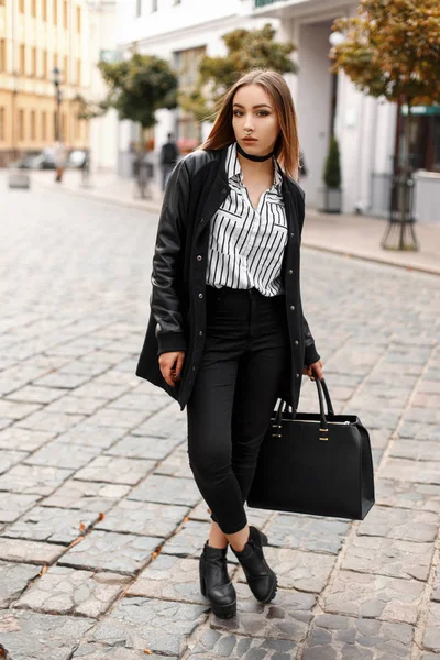 Jeune femme avec un nez percé dans une veste de printemps dans un chemisier rayé en jeans noirs en chaussures en cuir avec un sac à main noir se promène dans la ville près des bâtiments vintage dans le jour du printemps. Adolescente . — Photo