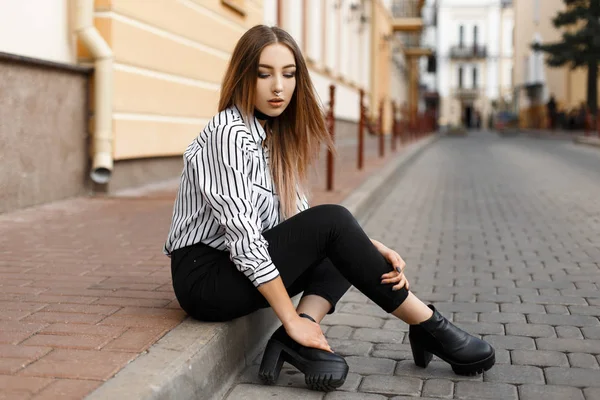 Moderna e elegante jovem mulher em uma blusa na moda em jeans preto em sapatos de couro está descansando sentado na rua perto de um vintage building.Pretty menina europeia desfrutando de dia de verão ao ar livre — Fotografia de Stock