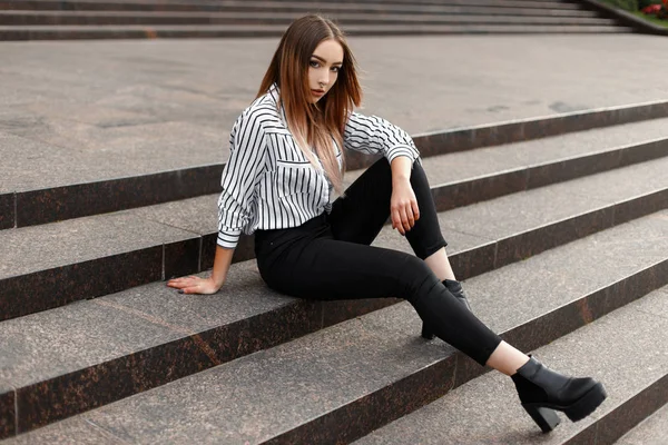 Joven mujer moderna en jeans de moda en una camisa a rayas de moda en zapatos de cuero negro posando en una escalera vintage en la ciudad en un cálido día de primavera al aire libre. Bastante adolescente en un paseo . —  Fotos de Stock