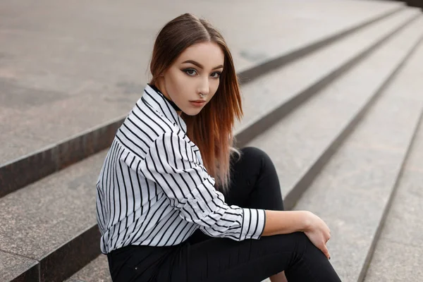 Jovem americana bonita em jeans na moda em uma blusa listrada preta e branca vintage com colares de veludo elegantes se senta nos degraus vintage da cidade em um dia quente de primavera. Menina bonita . — Fotografia de Stock