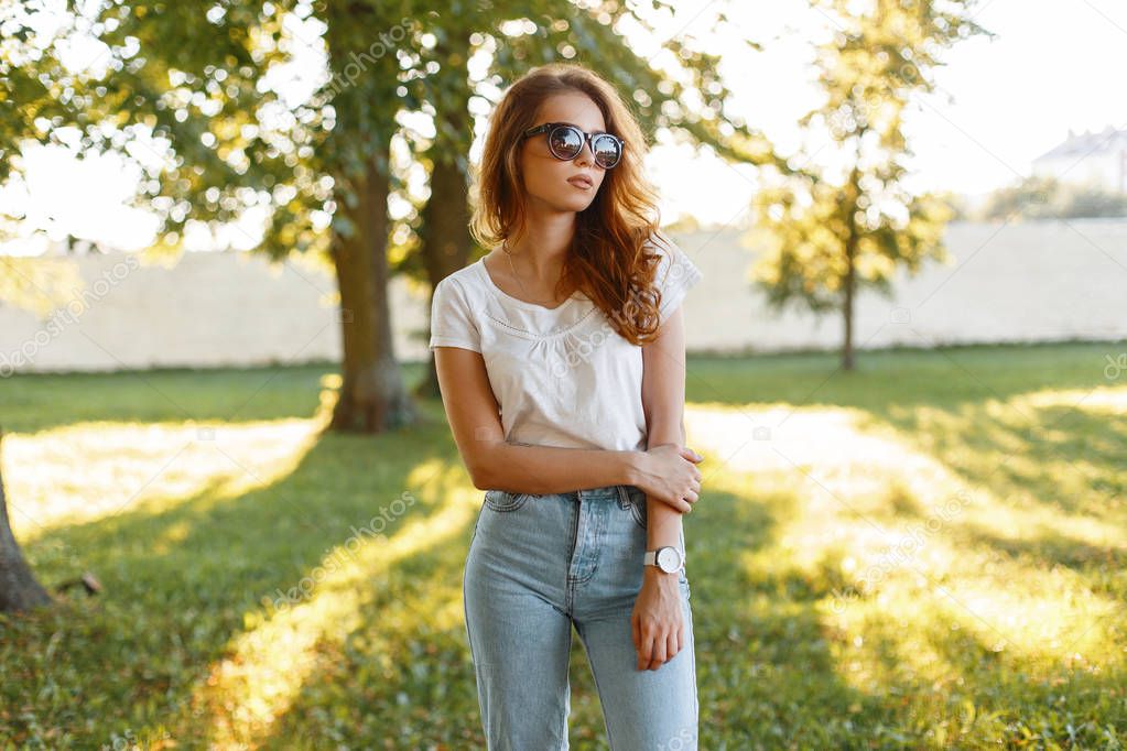 Beautiful redhead young hipster woman in vintage blue jeans in a white t-shirt in trendy sunglasses stands in a park among green trees on a summer sunset background. Cute girl enjoys a walk.