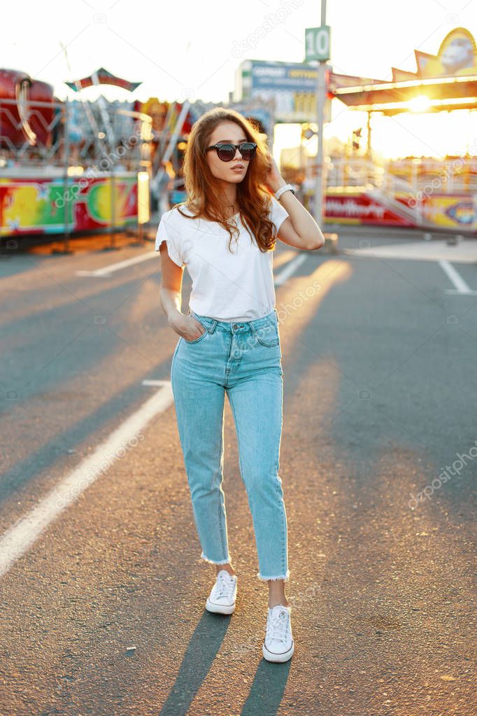 Young hipster woman in stylish T-shirt in trendy sunglasses in blue jeans in sneakers posing in an amusement park on the background of a bright orange summer sunset. Pretty girl enjoying the weekend.