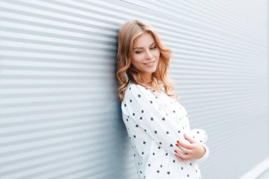 Beyaz moda kazak sarı saçlı Sevimli neşeli Avrupa şık genç kadın şehrin modern metal duvarın yakınında duruyor. Çekici neşeli kız model dinlenme açık havada.