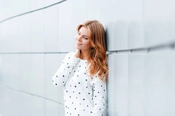 Cute european positive young woman in fashionable clothes is standing and smiling near the white modern wall indoors. Stylish positive girl model on vacation. — Stock Photo, Image