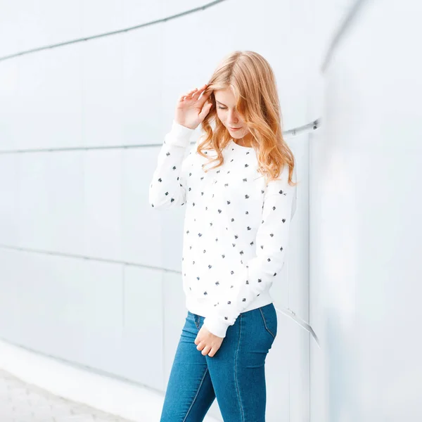 Веселая привлекательная молодая женщина с модной прической в белом свитере в старинных модных джинсах у современной стены города. Счастливая девушка-модель наслаждается выходными . — стоковое фото