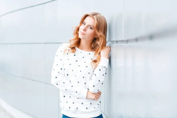 Ziemlich elegante positive junge blonde Frau mit einem schönen Lächeln in einem modischen Pullover in der Nähe einer weißen modernen Wand drinnen. stilvolle positive Mädchen-Modell im Urlaub. Jugendkleidung im Frühjahrsstil. — Stockfoto