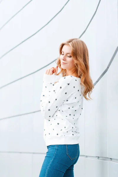 Fashionable modern young woman with a stylish hairstyle in a white sweater in blue trendy jeans near a modern wall in the city in the spring warm day. Cute happy girl model enjoys the weekend. — Stock Photo, Image