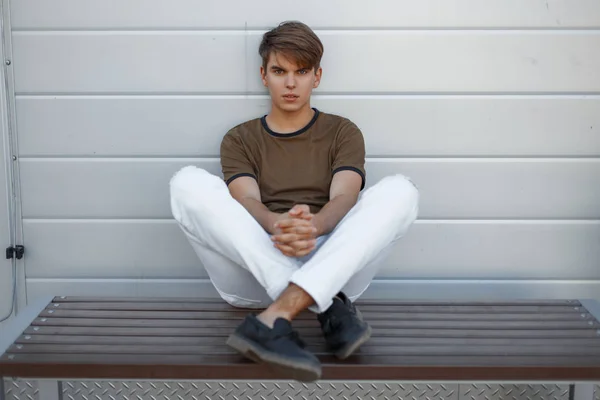 Hombre joven guapo americano en camiseta verde de moda en pantalones elegantes blancos en zapatos de cuero negro de moda se relaja sentado en un banco de madera cerca de una pared de metal vintage. Lindo chico moderno está descansando . — Foto de Stock