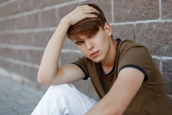 Jeune homme européen dans un T-shirt vert à la mode dans un pantalon blanc élégant avec une coiffure à la mode se détend assis près d'un mur de briques vintage. mignon moderne modèle de mode guy est au repos . — Photo