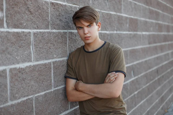 Cute handsome young man in a green t-shirt with a fashionable hairstyle posing standing near a vintage brick wall. American attractive guy model resting. — Stock Photo, Image